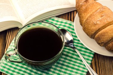 Kruvasan, açık kitap, ahşap masa peçeteyle üzerinde siyah kahve.