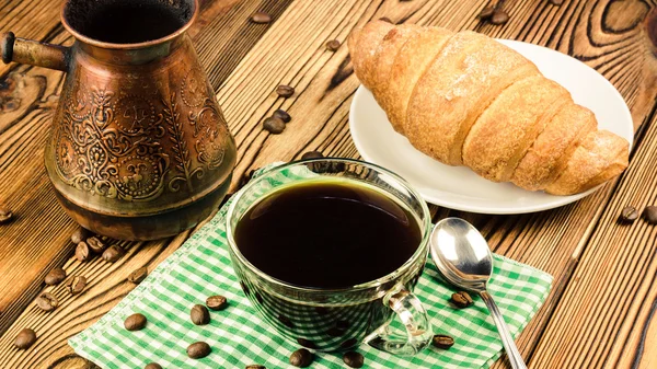 Taza de café negro en servilleta verde con croissant, cafetera turca, mesa de madera en la cafetería —  Fotos de Stock