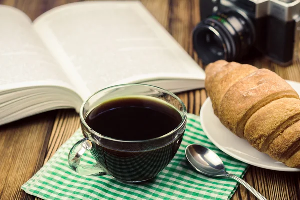 Taza de café negro en servilleta con croissant, libro abierto, cámara vintage, mesa de madera, concepto de viaje — Foto de Stock