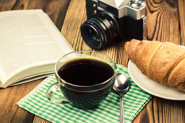 Taza de café negro en servilleta con croissant, libro abierto, cafetera turca, cámara vintage, mesa de madera — Foto de Stock