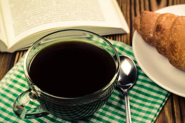 Чашка чорної кави на серветці з круасаном, відкрита книга, дерев'яний стіл . — стокове фото