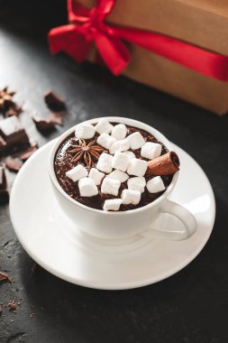 Beyaz bardakta sıcak çikolata, marşmelov ve karanlık arka planda noel hediyesi kutusu..
