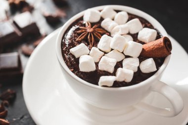 Beyaz bardakta sıcak çikolata, marshmallow, tarçın çubuğu, kırık çikolata küpleri ve yıldız anason, yakın çekim.