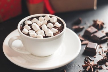 Beyaz bardakta sıcak çikolata, marşmelov ve karanlık arka planda noel hediyesi kutusu..
