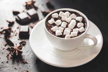 Beyaz bardakta sıcak çikolata, marşmelov, kırık çikolata küpleri ve siyah arka planda yıldız anason..