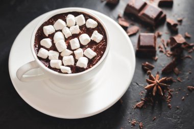 Beyaz bardakta sıcak çikolata, marşmelov, kırık çikolata küpleri ve siyah arka planda yıldız anason..