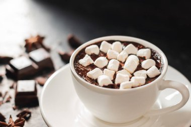 Beyaz bardakta sıcak çikolata, marşmelov, kırık çikolata küpleri ve siyah arka planda yıldız anason..