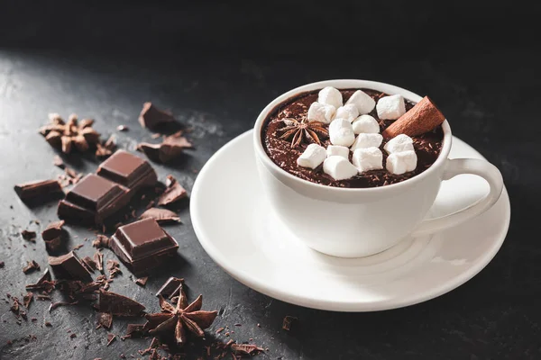 Hot chocolate drink in white cup with marshmallow, cinnamon stick, broken chocolate cubes and star anise on dark background.