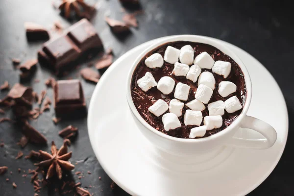 Hot chocolate drink in white cup with marshmallow, broken chocolate cubes and star anise on dark background.