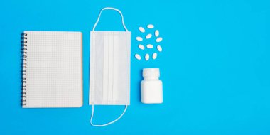 Beyaz tıbbi maske, ilaç şişesi ve mavi arka planda not defteri. Korunma, önleme ve tedavi Coronavirus, covid 19, grip. en üstteki görünüm.