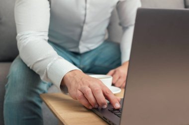 Tanınmayan bir adam, iş adamı ya da serbest çalışan biri kanepeye oturmuş, elinde kahve fincanıyla dizüstü bilgisayarda çalışıyor, evden çalışıyor..