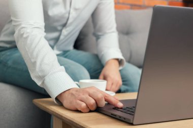 Tanınmayan bir adam, iş adamı ya da serbest çalışan biri kanepeye oturmuş, elinde kahve fincanıyla dizüstü bilgisayarda çalışıyor, evden çalışıyor..