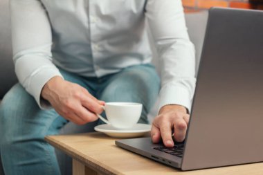 Tanınmayan bir adam, iş adamı ya da serbest çalışan biri kanepeye oturmuş, elinde kahve fincanıyla dizüstü bilgisayarda çalışıyor, evden çalışıyor..