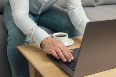 Tanınmayan bir adam, iş adamı ya da serbest çalışan biri kanepeye oturmuş, elinde kahve fincanıyla dizüstü bilgisayarda çalışıyor, evden çalışıyor..