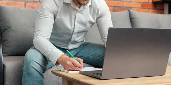 Unrecognized bearded man, businessman or freelancer sitting on a sofa and working on laptop from home, modern interior loft design.