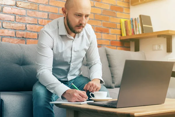 Bearded bald man, businessman or freelancer sitting on a sofa with pen and notepad in hand, working on laptop from home, modern interior loft design.