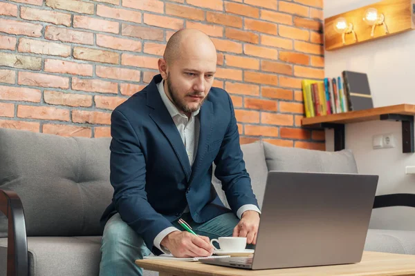 Bearded bald man, businessman or freelancer sitting on a sofa with pen and notepad in hand, working on laptop from home, modern interior loft design.