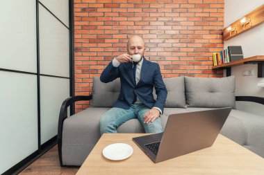 Çekici sakallı kel adam kanepeye oturup kahve içiyor, dizüstü bilgisayarına bakıyor, modern iç dekorasyon..