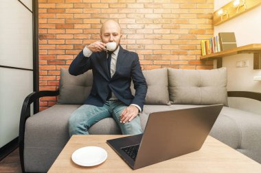 Çekici sakallı kel adam kanepeye oturup kahve içiyor, dizüstü bilgisayarına bakıyor, modern iç dekorasyon..