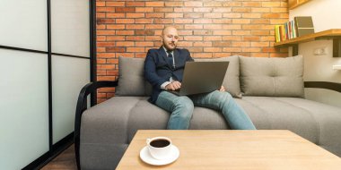 Kel sakallı adam, iş adamı ya da serbest çalışan. Kanepede oturuyor ve bilgisayarla evden çalışıyor. Modern iç mekan tasarımı..