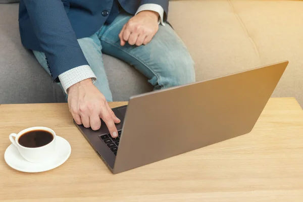 Unrecognized man, businessman or freelancer sitting on sofa and working at laptop on table with coffee cup, work from home.