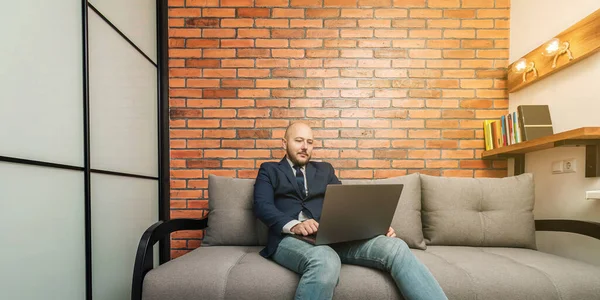 Bearded bald man, businessman or freelancer sitting on sofa and working on laptop from home, modern interior loft design.