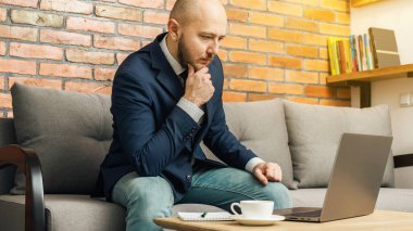 Kel sakallı adam, iş adamı ya da serbest çalışan. Kanepede oturuyor ve bilgisayarla evden çalışıyor. Modern iç mekan tasarımı..