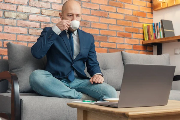 Attractive bearded bald man sitting on sofa and drinking coffee, looking at laptop, modern interior loft design.