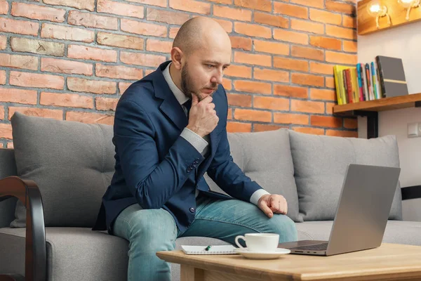 Bearded bald man, businessman or freelancer sitting on sofa and working on laptop from home, modern interior loft design.