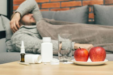 Evdeki tedavi virüsü enfeksiyonu, koronavirüs, covid-19 ya da grip, ilaç, ilaç, su, elma içeren masa, kanepede yatan hasta adam, hasta ve kendini kötü hissediyor..