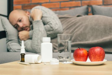 Evdeki tedavi virüsü enfeksiyonu, koronavirüs, covid-19 ya da grip, ilaç, ilaç, su, elma içeren masa, kanepede yatan hasta adam, hasta ve kendini kötü hissediyor..