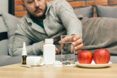 Evdeki tedavi virüsü enfeksiyonu, koronavirüs, covid-19 ya da grip, ilaç, hap, elma dolu bir masa ve kanepede yatan hasta adam bardağı suyla birlikte alıyor..
