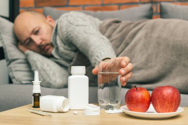 Evdeki tedavi virüsü enfeksiyonu, koronavirüs, covid-19 ya da grip, ilaç, hap, elma dolu bir masa ve kanepede yatan hasta adam bardağı suyla birlikte alıyor..
