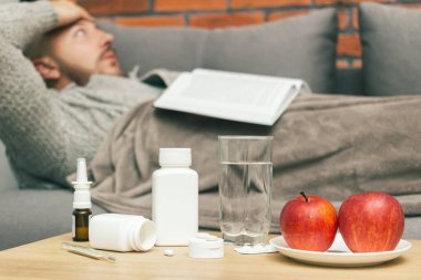 Evdeki tedavi virüsü enfeksiyonu, koronavirüs, covid-19 ya da grip, ilaçlı masa, haplar, elmalar odadaki, kitapsız arka plan hastası adam koltukta yatıyor..