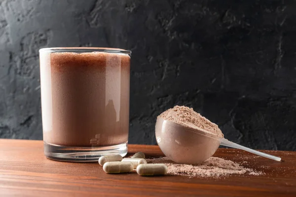 Glass with milkshake of whey protein isolate, protein powder in scoop, white capsules of amino acids and creatine, bodybuilding food supplements on wooden table and dark background.