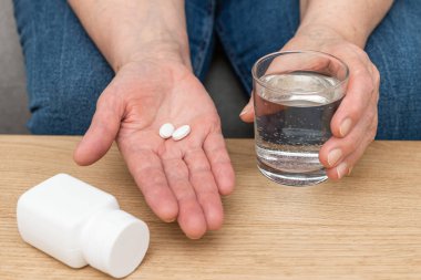 Yaşlı kadın avuçlarında tedavi için beyaz ağrı kesici ya da antibiyotik hapları, su dolu bardak, ilaç ve vitamin takviyesi konsepti, yakın çekim görüntüsü.