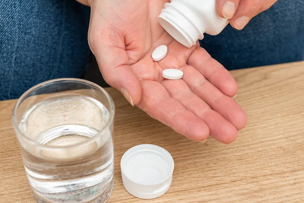 Yaşlı kadın avuçlarında tedavi için beyaz ağrı kesici ya da antibiyotik hapları, su dolu bardak, ilaç ve vitamin takviyesi konsepti, yakın çekim görüntüsü.