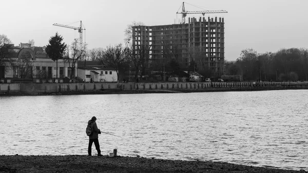 Fisherman standing on edge of shore with fishing rod near river in city, black and white — Stock Photo, Image