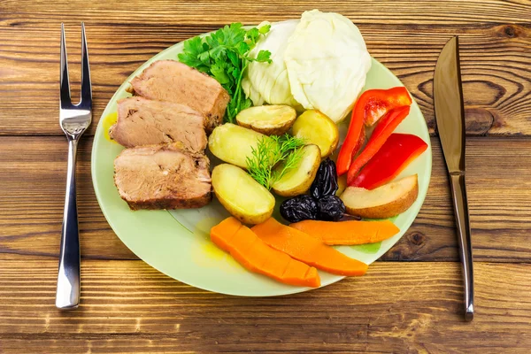 Comida saludable, carne de cerdo en rodajas con varias verduras guisadas en plato, cuchillo y tenedor sobre fondo de madera — Foto de Stock