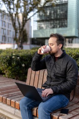 Üniversitede bankta oturan genç adam internette çalışıyor ve kahve içiyor.