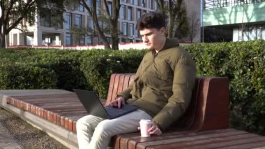 Üniversitede güneşin altında oturan genç bir adam internetten kahve içiyor.