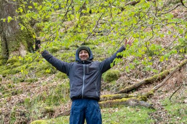 Şapkalı bir adam gülümseyerek ormanda kollarını kaldırıyor.