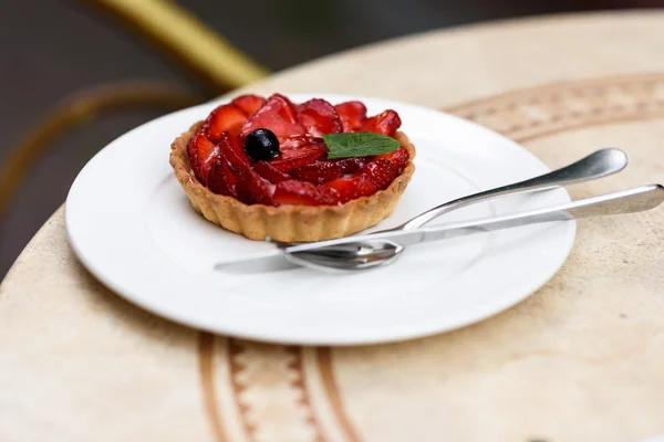 Foto de un pastel de frutas con fresas, menta y grosella negra, en un plato blanco con cuchillo y tenedor en una mesa de restaurante . — Foto de Stock