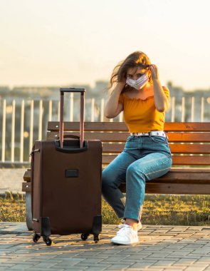 Park yerinde bavullu yüz maskesi takan bir kız. Salgın sırasında seyahat