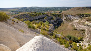 Büyüyen çimenler ve çalılarla kireçtaşı ocağı, beyaz yamaçlar, köy, tepeler ve uzaktaki tarlalar görünür.