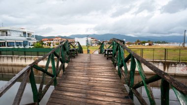 Su kanalının üzerindeki ahşap köprü, uzakta evler ve tepeler, bulutlu gökyüzü, Stavros, Yunanistan
