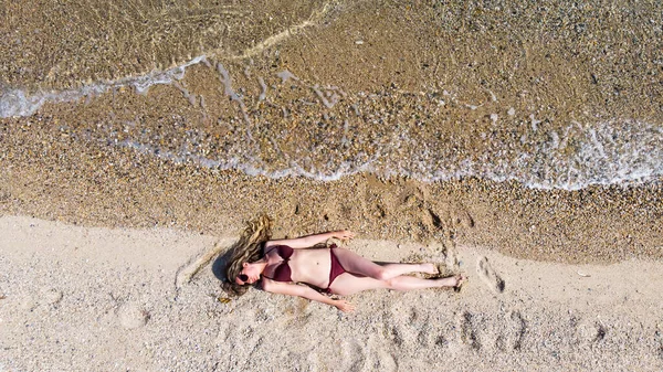 一个戴着太阳镜的高加索金发女人躺在海滩上 海浪冲向身体 身穿勃艮第泳衣 — 图库照片