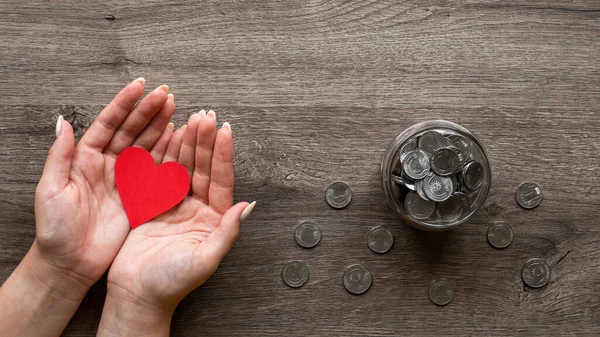 Woman is holding a red hearth, the can with metal coins inside and around it. Woman hands. Wooden background. Top view