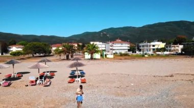 Siyah şapkalı ve güneş gözlüklü bir adam Ege sahilinde güneş yataklı ve şemsiyeli bir dron fırlatıyor. Asprovalta Panoraması 'nda binalar, yeşillikler, yeşil tepeler. Güneşli bir gün. Yunanistan