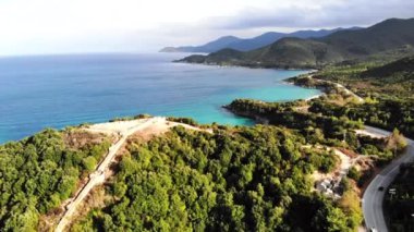 Yeşillikle kaplı tepeler. Tepenin tepesindeki antik yapıların kalıntıları. Ege kıyısı. Hareket halindeki arabalarla yol. Yunanistan